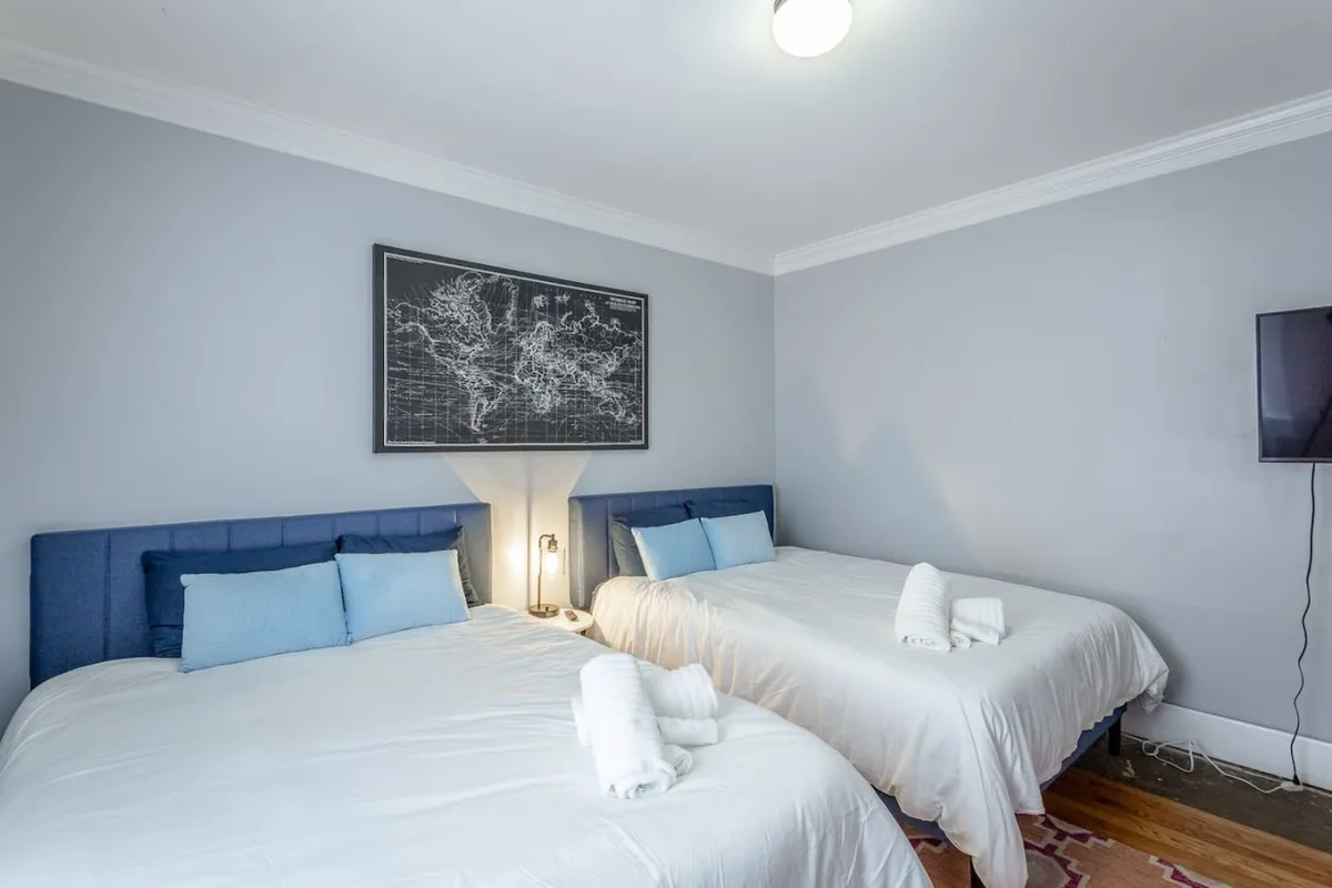 A bright and inviting bedroom featuring two beds with blue headboards, soft white linens, and world map artwork above, illuminated by warm bedside lamps. The overall ambiance is both comfortable and serene, ideal for restful nights or morning chats.
