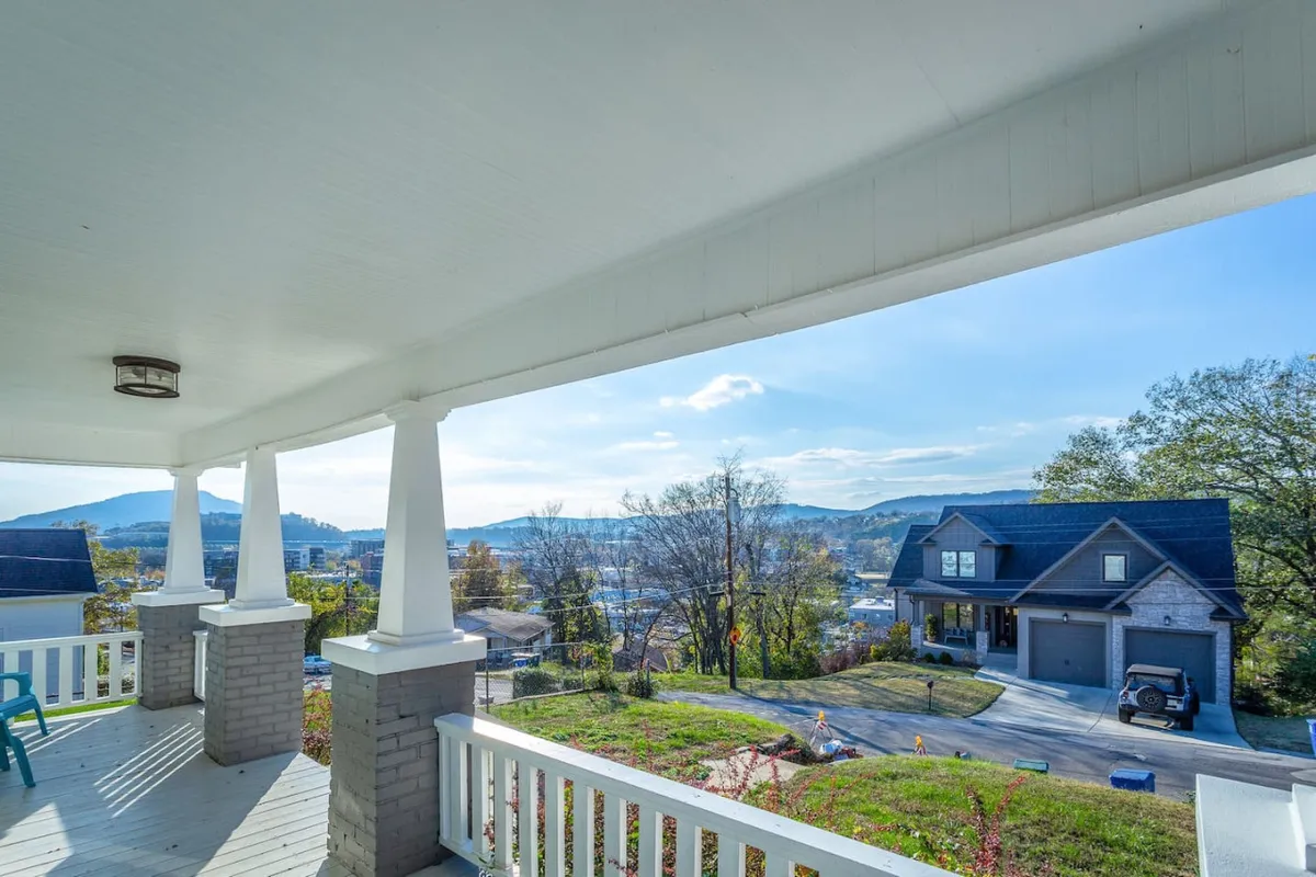 A welcoming front porch with classic columns, a blue chair, and a picturesque mountain view. The porch ambiance is serene, suggesting relaxation and socializing after a day exploring the city. The neighboring house is visible, underscoring the neighborhood setting.