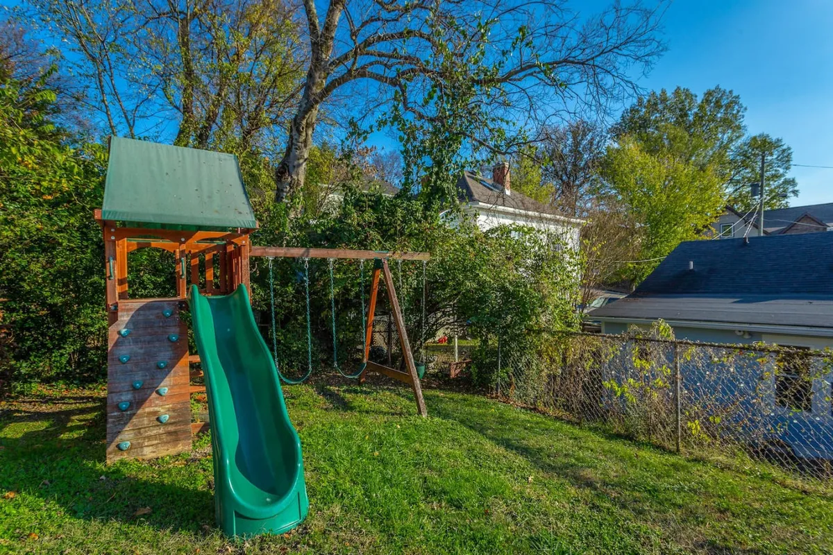 A cheerful, tree-lined playground featuring swings, a slide, and bright green grass. Children play and families gather for picnics or relaxation in an inviting natural setting. The open spaces and equipment make it ideal for group activities and family fun outdoors.