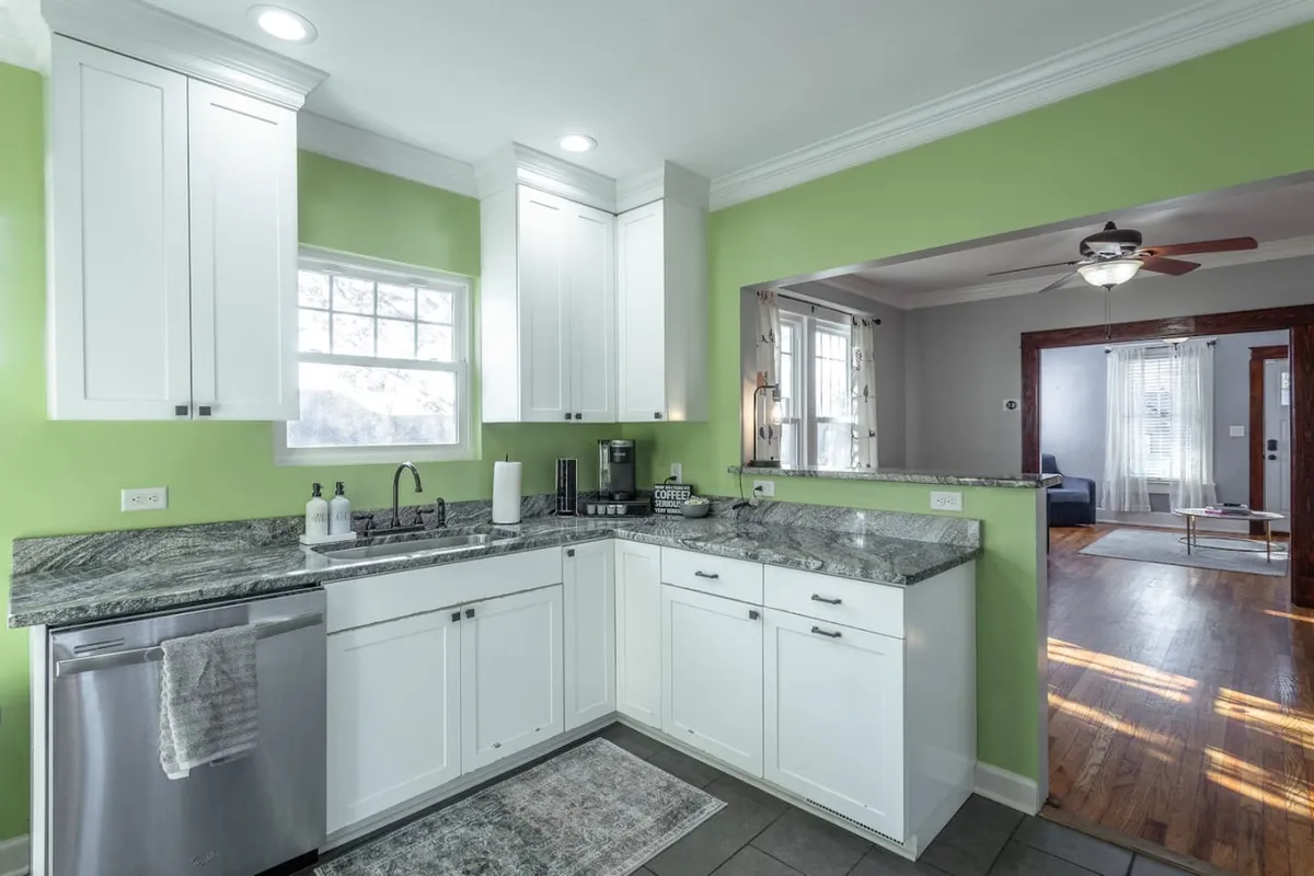 A cheerful kitchen scene with green walls, granite countertops, and white cabinetry. Kitchen appliances are visible, and the space is filled with bright, natural light, suggesting a lively and inviting environment perfect for cooking and socializing.