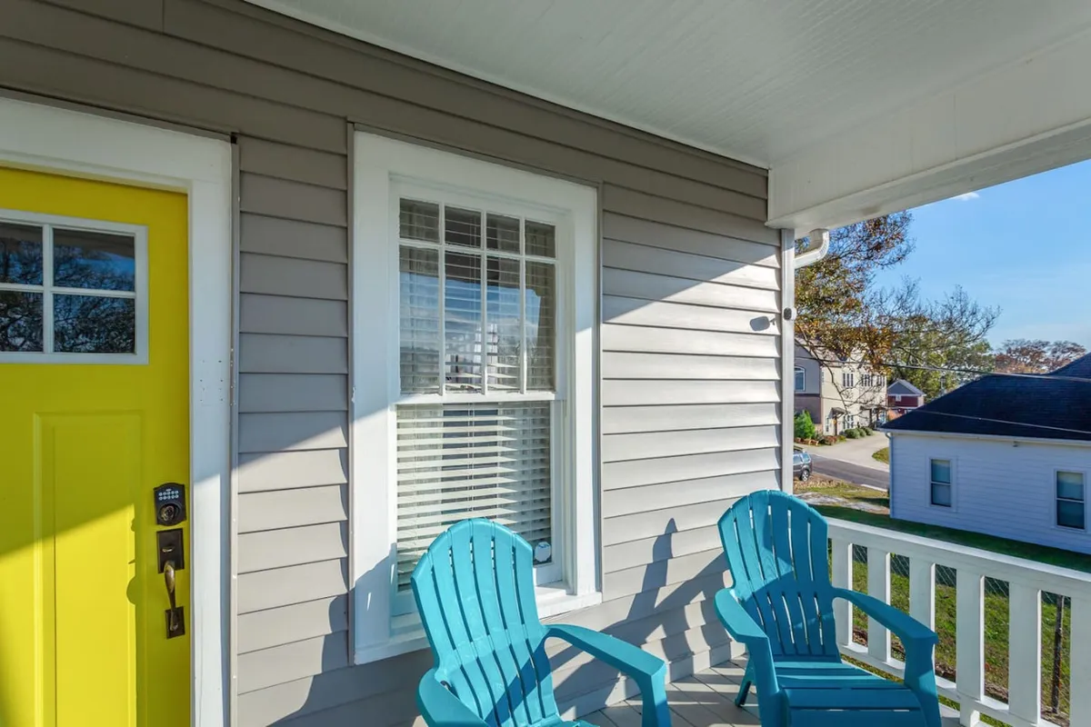 A relaxed front porch scene with a yellow front door, blue Adirondack chairs, and white railing. The porch exudes comfort and encourages quiet moments or casual group discussions, with ample natural light and a welcoming vibe, reflecting the flexibility of using different home spaces for collaboration or downtime.