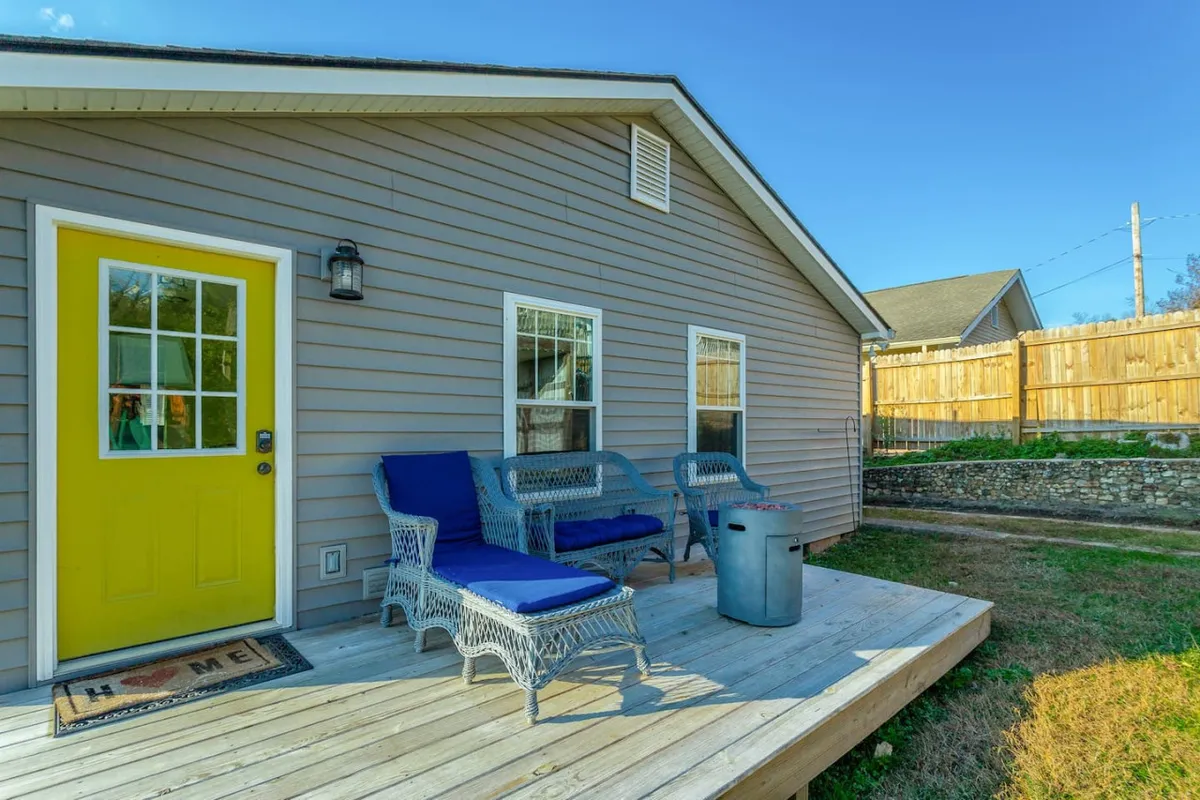 A welcoming exterior shot of a home featuring a bright yellow front door, a cozy blue seating area, and a wooden deck. The setup is inviting, with the deck providing a comfortable space for groups to relax, socialize, or transition between indoor and outdoor activities. The lush greenery and natural light set the tone for both work and leisure moments just outside the door.